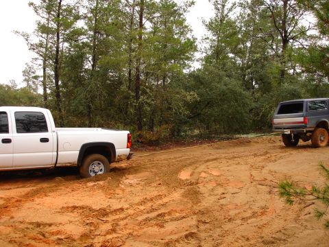 1989 Ford Bronco - A white pickup truck is stuck in the mud while a dark SUV attempts to tow it out.