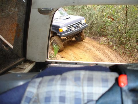 1989 Ford Bronco - A dark blue Ford Bronco navigates a sandy, wooded trail with its front wheels turned.