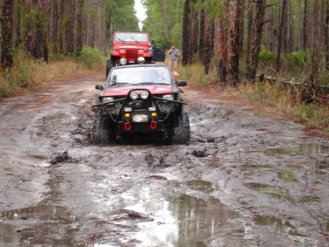 1989 Ford Bronco - Two off-road vehicles navigate a muddy trail through a forest.