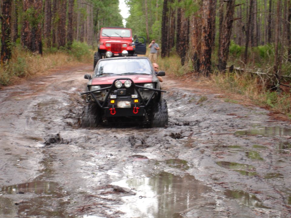 Red off-road SUV with winch and auxiliary lights driving through mud, with a red Jeep behind it