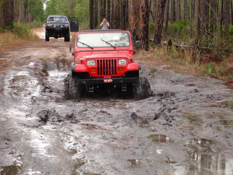 1989 Ford Bronco - Two off-road vehicles navigate a challenging muddy path in a wooded area.
