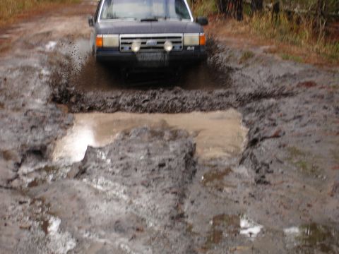 1989 Ford Bronco - A dark SUV splashes through a large mud puddle on a dirt road.