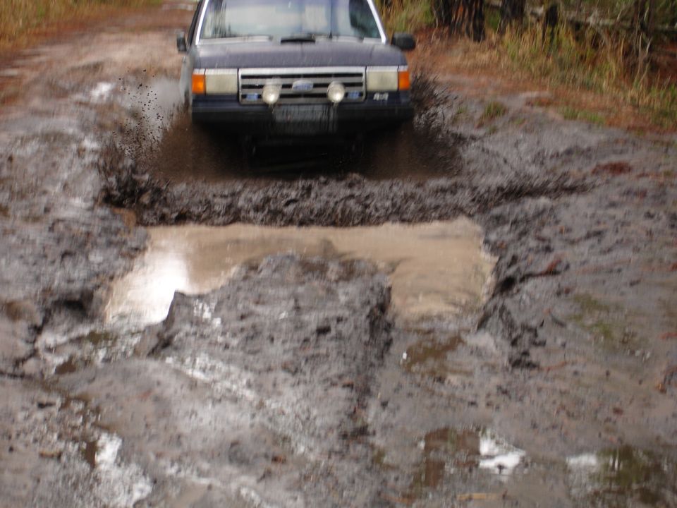 Dark SUV with a prominent grille and dual headlights navigating a deep mud puddle