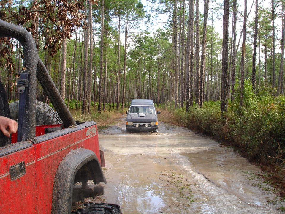 Red off-road vehicle in foreground, gray van driving through mud in forest