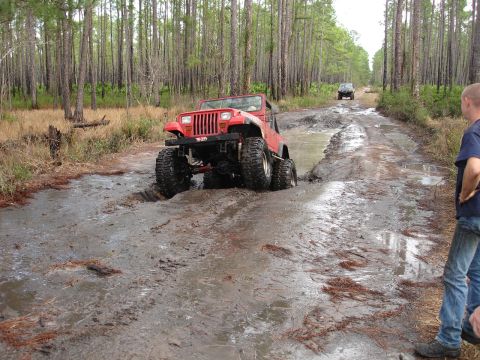 1989 Ford Bronco - A red Jeep navigates a challenging muddy path in a wooded area, with another vehicle visible in the distance.