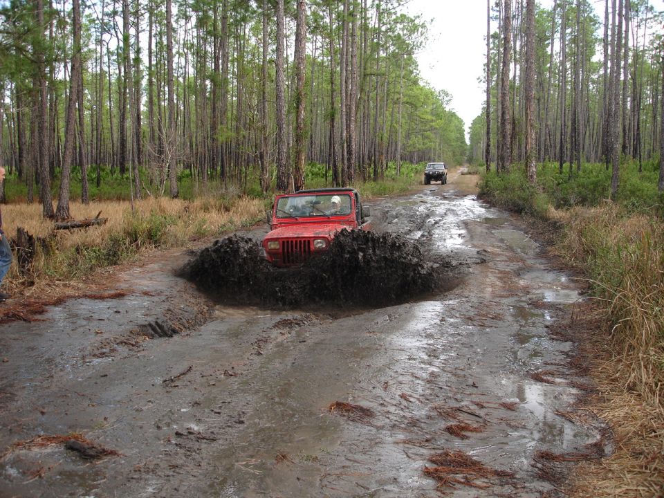 Red Jeep Wrangler driving through a deep mud puddle on a dirt road in a forest. Another SUV is visible further down the road