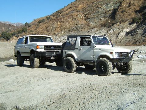 1988 Ford Bronco - Two off-road vehicles, a Ford Bronco and a Suzuki Samurai, are parked side-by-side on a dirt track with a rocky hillside behind them.