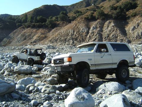 1988 Ford Bronco - Two off-road vehicles navigate a rocky riverbed with a steep, vegetated hillside in the background.
