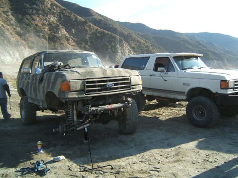1988 Ford Bronco - Two Ford Broncos are parked on a beach, one undergoing maintenance with its front wheel removed.