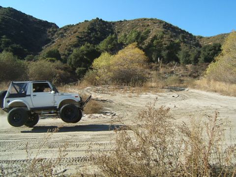 1988 Ford Bronco - Suzuki Samurai jumps a dirt track in a hilly, arid landscape.