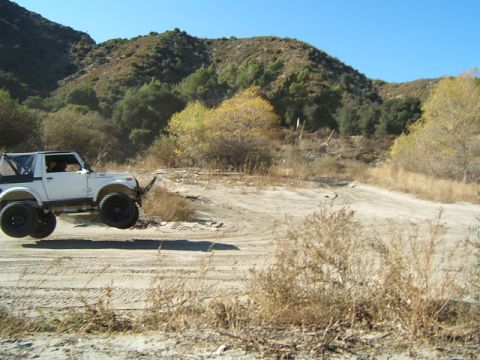 1988 Ford Bronco - A white off-road vehicle is captured mid-air on a dusty trail, with dry vegetation and hills under a clear blue sky.