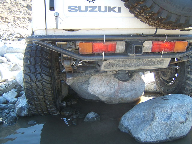 White Suzuki Samurai rear with large tires, stuck on rocks in water