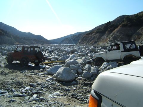 1988 Ford Bronco - Two off-road vehicles navigate a rocky riverbed, with mountains visible in the distance under a clear sky.