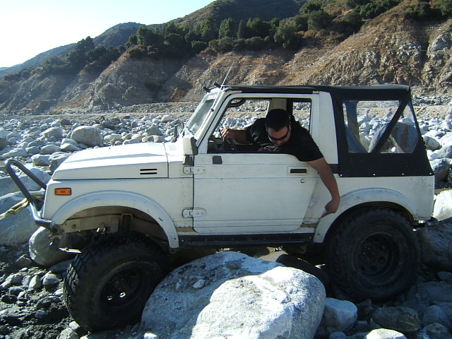 White off-road SUV with a black soft top navigating a rocky terrain. Driver is leaning out the window
