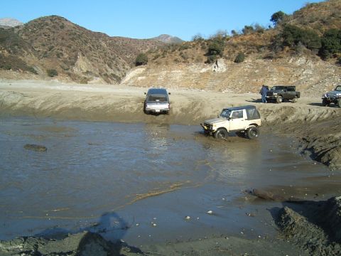 1988 Ford Bronco - Multiple off-road vehicles traverse a muddy, water-filled landscape under a clear blue sky.
