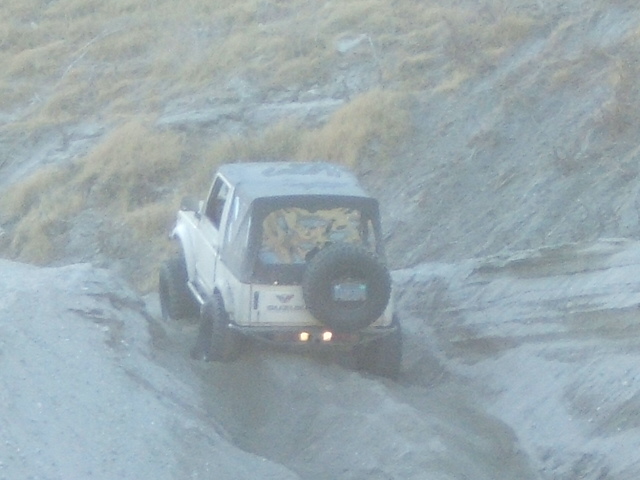 Suzuki Samurai with soft top driving up a dirt hill, spare tire mounted on the back