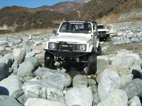 1988 Ford Bronco - Two off-road vehicles navigate a rocky riverbed under a clear blue sky.