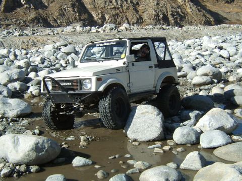 1988 Ford Bronco - A white Suzuki Samurai navigates a rocky riverbed, its tires gripping the uneven terrain.