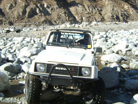 1988 Ford Bronco - A white Suzuki Samurai navigates a rocky terrain, with another white truck visible in the background.