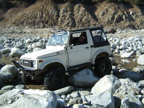 1988 Ford Bronco - A Suzuki Samurai navigates a rocky riverbed, with another off-road vehicle in the distance.