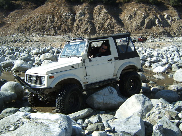 White Suzuki Samurai driving over rocks in a riverbed. Another vehicle is visible in the background
