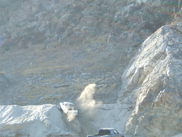 White pickup truck kicking up dust as it goes down a steep dirt hill. Another pickup truck is visible below