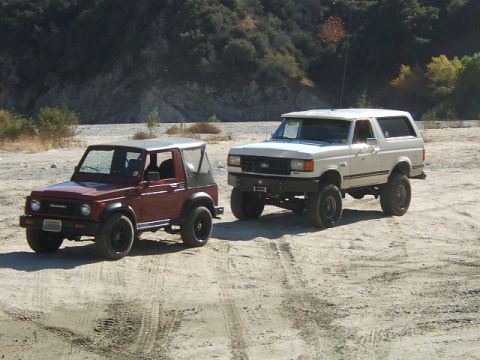 1988 Ford Bronco - Two off-road vehicles, a white Ford Bronco and a red Suzuki Samurai, are parked on a dirt road with hills in the background.