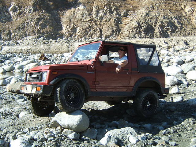 Red Suzuki Samurai with black soft top, driver visible, navigating a rocky, dry riverbed