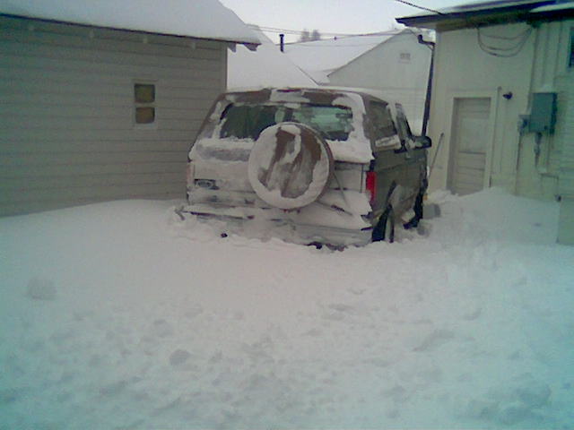 Dark full-size SUV with rear spare tire cover buried in deep snow between two buildings