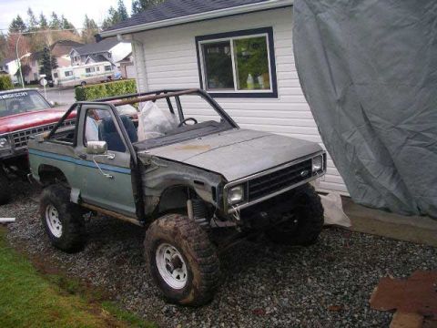 1984 Ford Bronco II - A light blue 1984 Ford Bronco II is parked next to a red vehicle, off-road tires and a roll cage.
