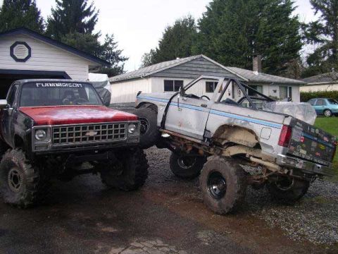 1984 Ford Bronco II - Two lifted trucks are parked outside a house. The front truck has a red hood and a large grille.