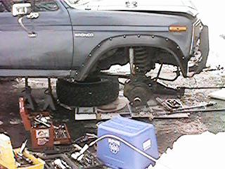1986 Ford Bronco - A gray 1986 Ford Bronco is shown from the side, with its front wheel removed and suspension components visible, surrounded by tools.