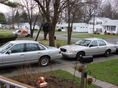 2003 Ford Crown Victoria - Two silver sedans are parked on a residential street, one in front of the other.