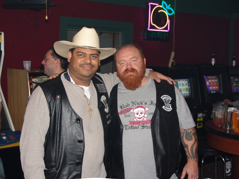 Two men posing in a bar, one in a cowboy hat, both wearing motorcycle club vests