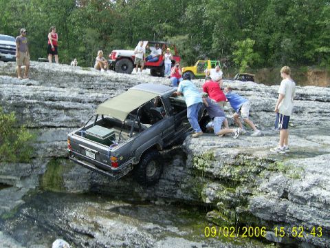 1979 Ford Bronco - Several people attempt to push a black SUV off a rocky ledge into the water below.