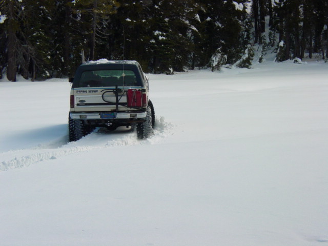 1986 Ford Bronco driving off-road in deep snow, with a spare tire and gas can mounted on the back