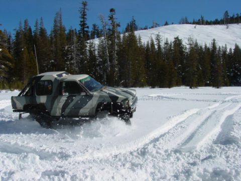 1986 Ford Bronco - A camouflaged pickup truck navigates a snowy landscape, kicking up snow as it drives through deep powder.