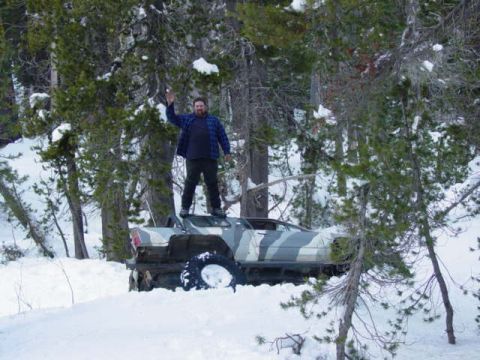 1986 Ford Bronco - A man stands atop a camouflaged SUV partially buried in snow, waving to the camera.