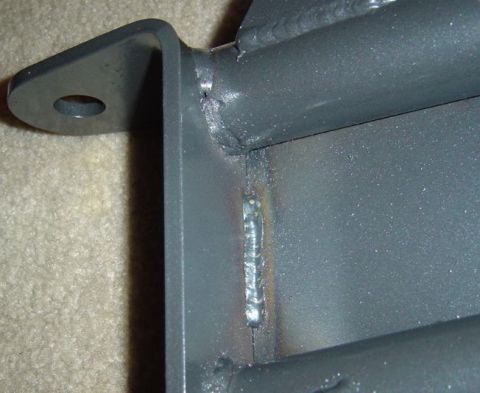 1986 Ford Bronco - Close-up of a gray-painted fabricated steel bracket photographed on carpet, showing a rough weld bead with visible slag and heat discolorat…