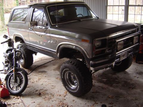 1986 Ford Bronco - A two-tone gray Ford Bronco full-size SUV lifted on large BFGoodrich All-Terrain T/A tires with chrome wheels, parked in a garage at a 3/4…