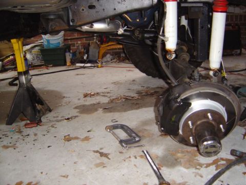 1986 Ford Bronco - Underside view of a vehicle's front suspension and brake assembly, showing an exposed drum/hub with wheel studs, two white coil-over shocks…