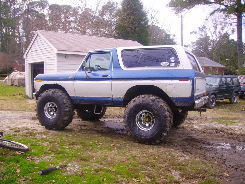 Blue and white Ford Bronco with large off-road tires parked on muddy ground near a garage