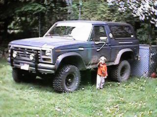 1986 Ford Bronco - A dark blue 1980s Ford Bronco is parked on grass with a child standing beside the driver's door. The vehicle has large off-road tires and a…