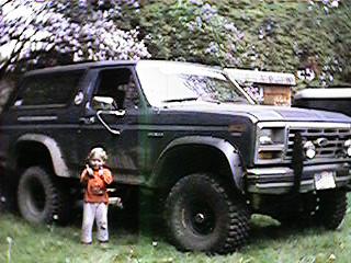 1986 Ford Bronco - A child stands next to a dark-colored Ford Bronco from the 1980s.
