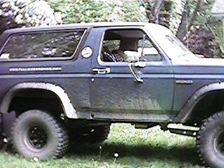 1986 Ford Bronco - A dark blue Ford Bronco is seen from the side, with off-road tires and some visible rust.