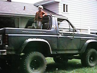 1986 Ford Bronco - Child stands in the open roof of a dark green 1980s Ford Bronco.