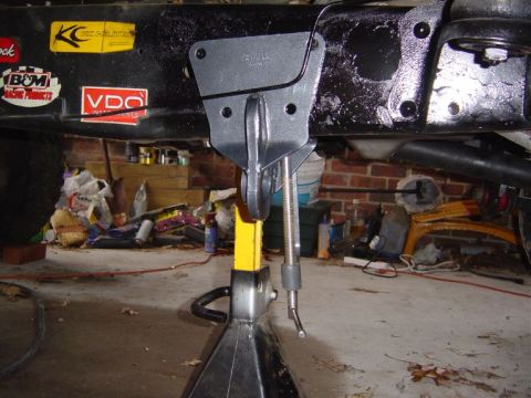1986 Ford Bronco - Underside view of a black vehicle frame with a shifter or linkage mockup being fitted, supported by a yellow jack stand, showing a threaded…