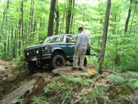 1986 Ford Bronco - A man stands beside a green Ford Bronco navigating a challenging off-road path through a dense forest.