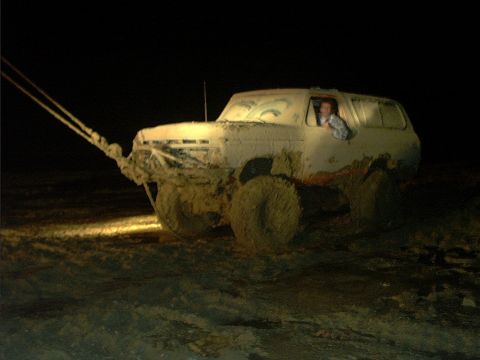 1978 Ford Bronco - Night shot of a lifted white full-size Ford Bronco caked in thick mud, being pulled by tow straps hooked to its front end while the driver…