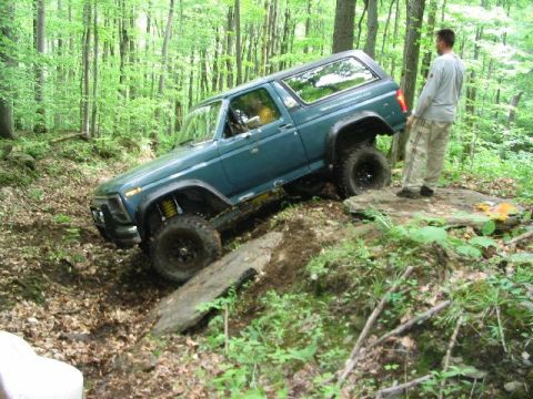 1986 Ford Bronco - This teal Ford Bronco navigates a challenging, rocky trail in a dense forest.